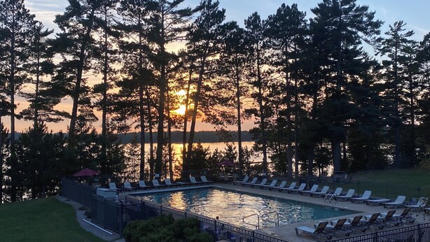 A poolside area with lounge chairs and a view of the sunset over a lake, surrounded by tall trees. - SUITE 206 W/ Garage@EAGLE WATERS RESORT (eagle river)
