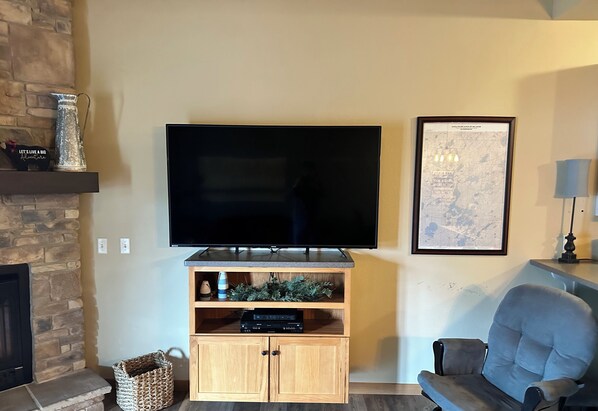 A living room with a flat-screen TV, wooden entertainment unit, a framed map, a fireplace, and a grey armchair.