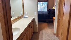 A bathroom with a wooden vanity, a large mirror, and a window leading to a bedroom with a bed and a ceiling fan.