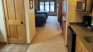 A narrow hallway leading to a bedroom with two beds, a kitchenette with a microwave, and a ceiling fan.