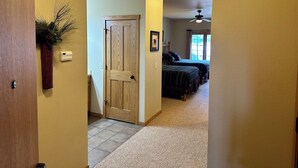 A hallway leading to a bedroom with a ceiling fan, a door with a decorative plant, and a window with curtains.