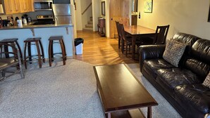 A modern kitchen with wooden cabinets, a stainless steel refrigerator, and a microwave. There is a dining area with a wooden table and chairs, and a black leather sofa with a patterned pillow. - Spirit of the Northwoods (eagle river)