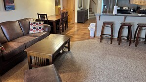 Un salon moderne avec un canapé en cuir, une table basse en bois et un coin repas comprenant des chaises et une table en bois. On aperçoit à l’arrière-plan une cuisine avec des meubles en bois et un réfrigérateur en acier inoxydable.