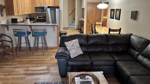 Een moderne woonkamer met een zwarte leren bank, een salontafel met afstandsbedieningen, en een keuken met houten kastjes en roestvrijstalen apparatuur.