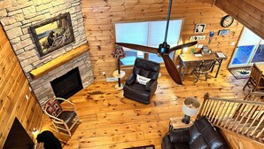 Interior acogedor de una cabaña con chimenea de piedra, mobiliario de cuero, ventilador de techo y suelo de madera rústica.