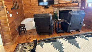 Una sala de estar acogedora con paredes de madera, una chimenea, dos sillones de cuero, un televisor y una mesita.