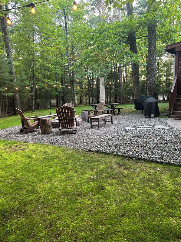 Un espacio al aire libre con sillas Adirondack, una chimenea y luces colgantes.