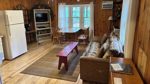 Una sala de estar acogedora con suelo de madera, un sofá, una mesa de comedor con sillas, un banco pequeño rojo, un televisor y un refrigerador.