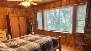 A wooden cabin bedroom with a bed, a chair, and a window with blinds. - Cabin 14A by Eagle Waters Resort (eagle river)