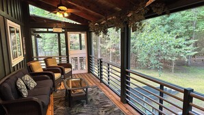 A wooden deck with wicker furniture, a glass table, and a view of the surrounding forest.