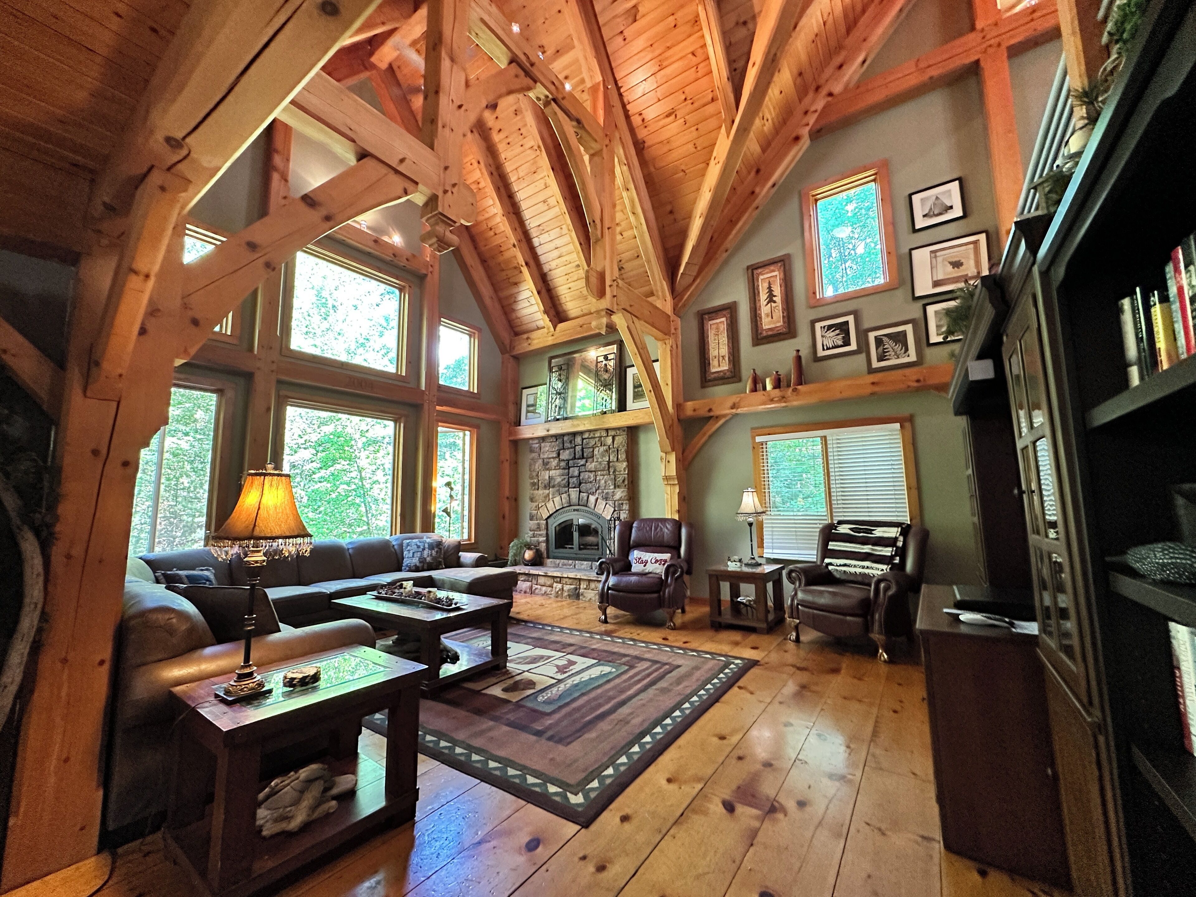 A cozy living room with a stone fireplace, wooden beams, large windows, and a fireplace mantel with framed pictures.
