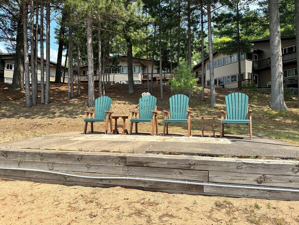 A row of Adirondack chairs and a small table on a concrete patio surrounded by sand and sparse grass, with a wooden fence in the foreground.