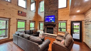A spacious living room with a stone fireplace, wooden walls, and large windows offering views of the outdoors.