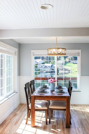 A dining area with a wooden table and chairs, a chandelier, and a view of a lake and houses through large windows. - Long Lake Retreat (Lacey)