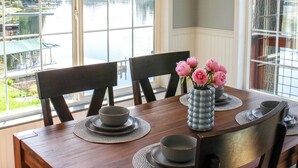 Une table à manger avec des chaises, un vase rempli de fleurs et une vue sur un lac, visible à travers de grandes fenêtres.