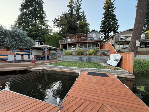A wooden dock extending over a pond, surrounded by a landscaped garden with various plants and a seating area with chairs. - Long Lake Retreat (Lacey)