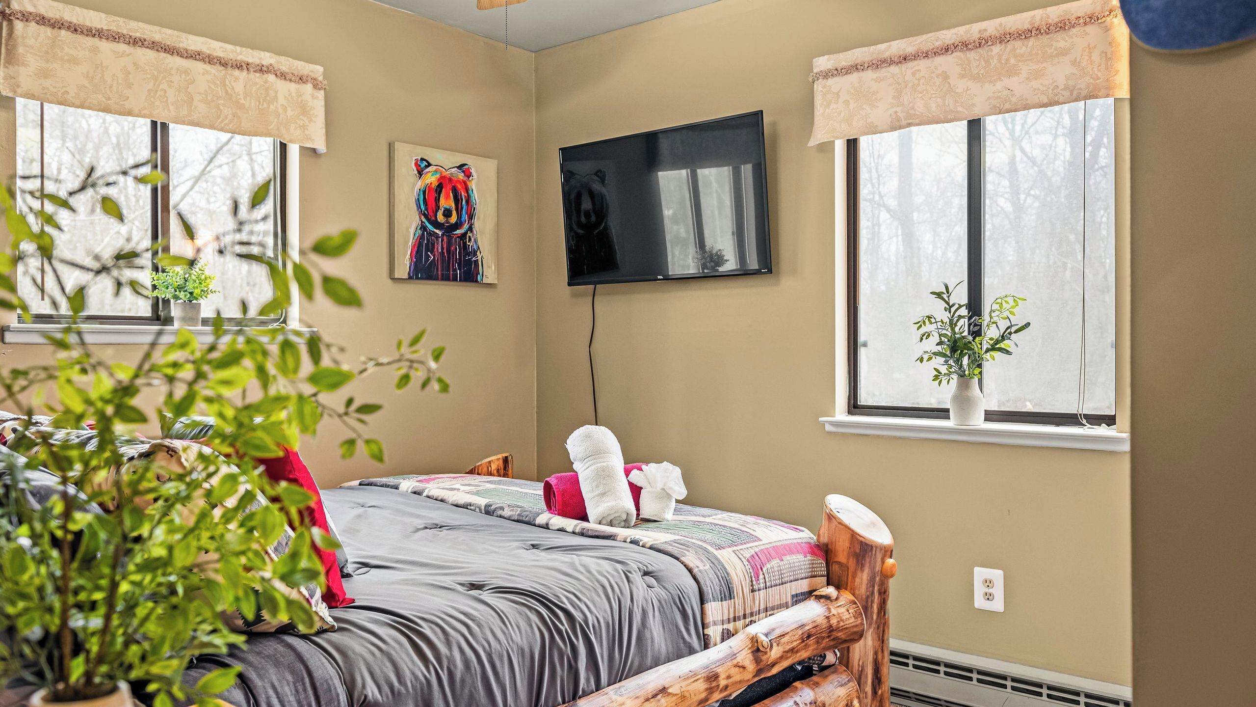 A bedroom with a bed, a TV, a window with a plant, and a painting on the wall.