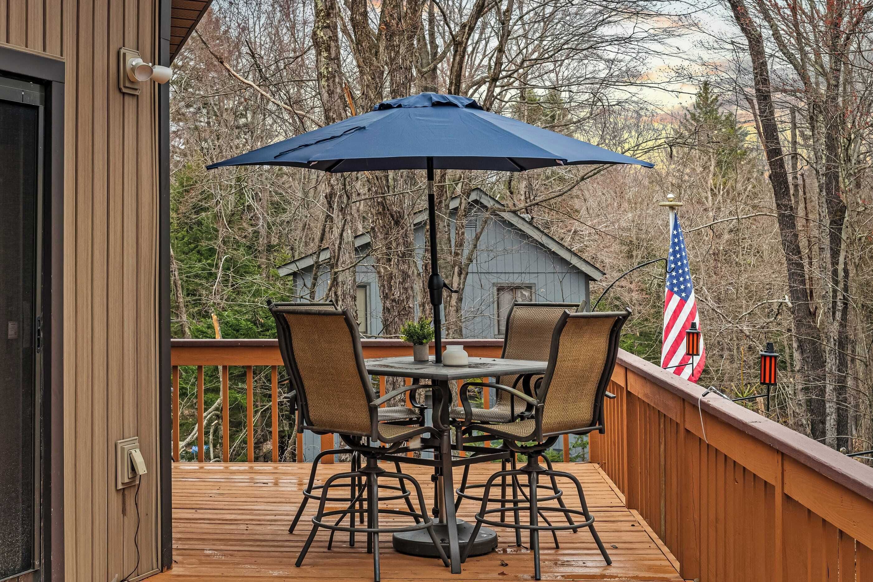 Eine Holzterrasse mit Tisch und Stühlen unter einem blauen Sonnenschirm, eine amerikanische Flagge und Blick auf Bäume.