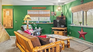 A cozy living room with a green wall, a wooden sofa, a TV, and a window with a view. - Atlas Point Lodge by Avantstay Hot Tub + Sauna + Kayaks Pet-friendly Cabin Lake Access (Pocono Lake)