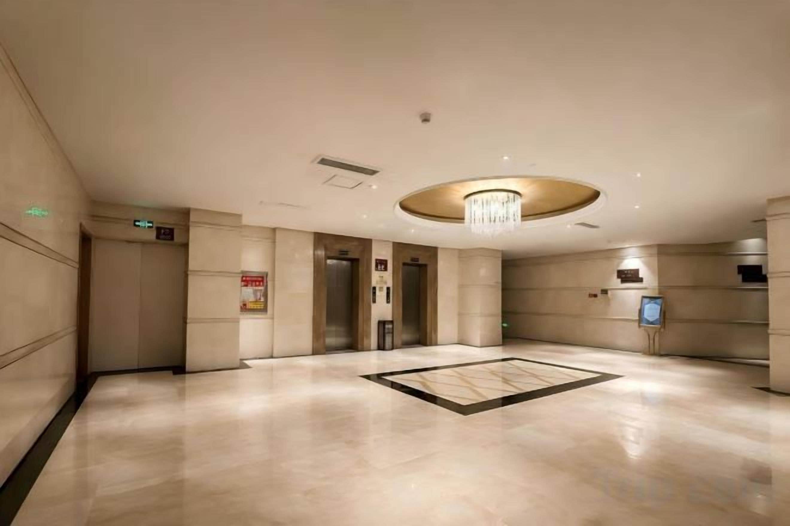 A spacious lobby with a central chandelier, elevators, and a reception desk.
