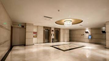 A spacious lobby with a central chandelier, elevators, and a reception desk.