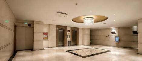 A spacious lobby with a central chandelier, elevators, and a reception desk.