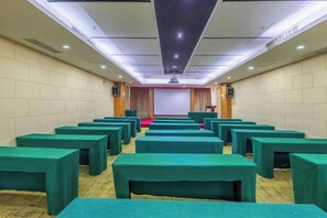 Un salón de conferencias con filas de mesas y sillas, un escenario y una pantalla de proyector.