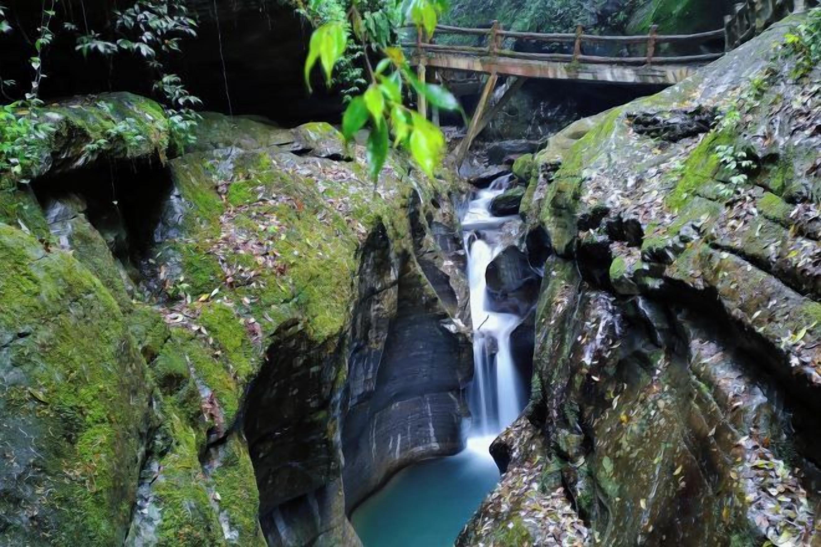 A natural waterfall with a wooden bridge, surrounded by moss-covered rocks and greenery.