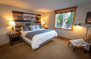 A bedroom with a large bed, two bedside tables with lamps, a window with a view of greenery, and a wooden headboard with a picture.