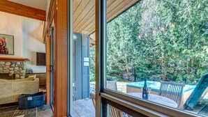 A cozy living room with a stone fireplace, a sofa, and a large window overlooking a backyard with a hot tub and wooden deck. - Large 3BD Village Townhome (Whistler)