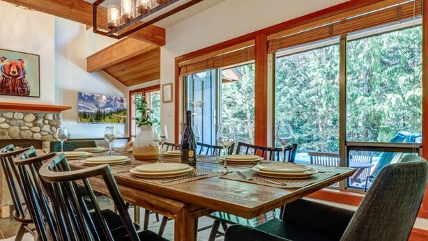 A dining area with a wooden table set for four, surrounded by chairs, and large windows offering a view of trees. - Large 3BD Village Townhome (Whistler)