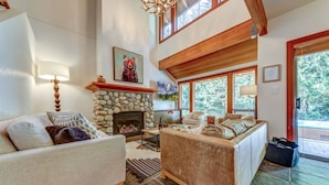 A spacious living room with a stone fireplace, a large sofa, a coffee table, and a view of the outdoors through large windows. - Large 3BD Village Townhome (Whistler)