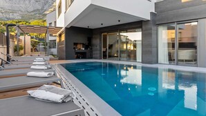 A modern pool area with lounge chairs and a clear blue pool, surrounded by a contemporary building with large windows and a stone facade. - Villa Mosaicus with Pool (Makarska)