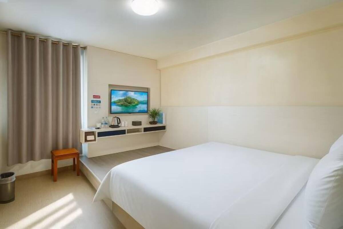 A hotel room with a bed, a TV mounted on the wall, a small table, and a window with curtains.