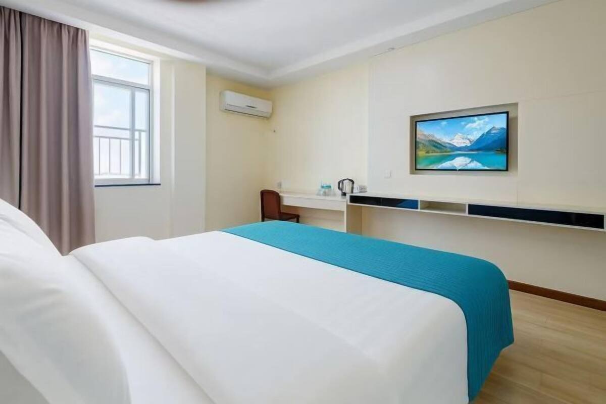 A hotel room with a bed, a desk, a chair, a window with curtains, an air conditioning unit, and a picture on the wall.