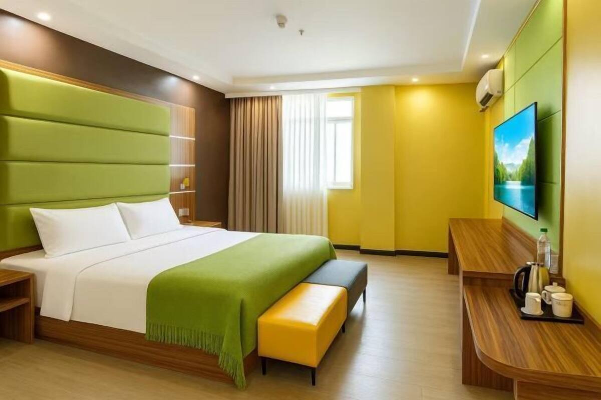 A hotel room with a large bed, a bench, a TV, and a desk with a kettle and cups.