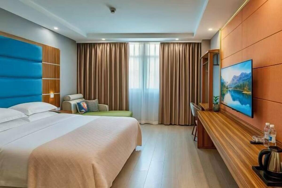 A hotel room with a bed, a desk, a chair, a sofa, a painting, and a window with curtains.