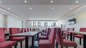 A spacious dining area with high tables and chairs, a flat-screen TV mounted on the wall, and large windows providing natural light.