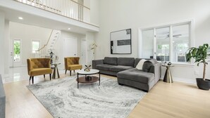 A spacious living room with a grey sectional sofa, two mustard-colored armchairs, a round coffee table, and a large grey rug. A staircase leads to an upper level, and a large window provides natural light.