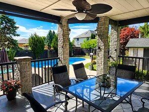 Un patio techado con una mesa y sillas de vidrio, un ventilador de techo y vistas a una piscina y casas.