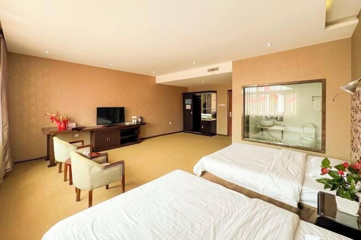 Quarto de hotel com duas camas, uma escrivaninha com televisão, um espelho e um espaço de estar com cadeiras.