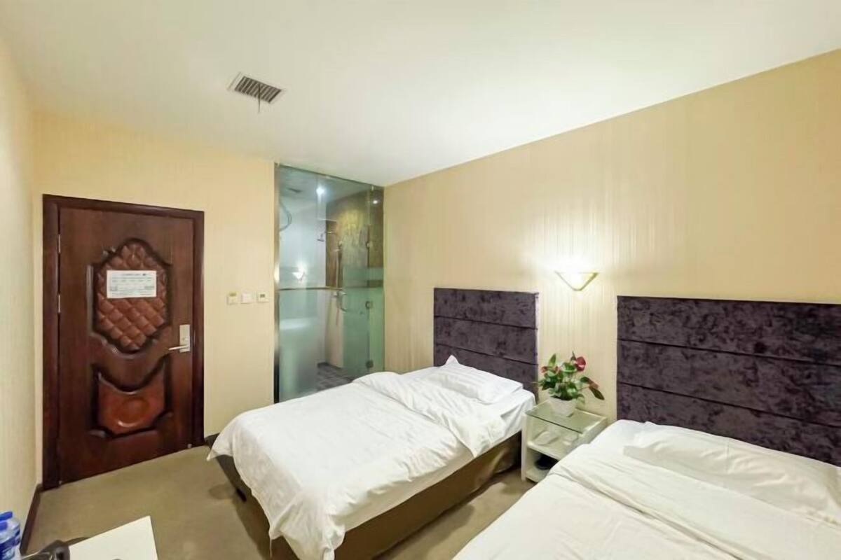 Quarto de hotel com duas camas, porta de madeira, área de chuveiro em vidro e mesa de cabeceira com um vaso de flores.