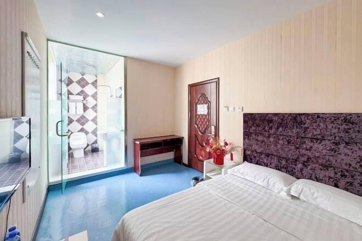 Quarto de hotel com cama, escrivaninha, cabine de hidromassagem em vidro e porta com detalhe decorativo.