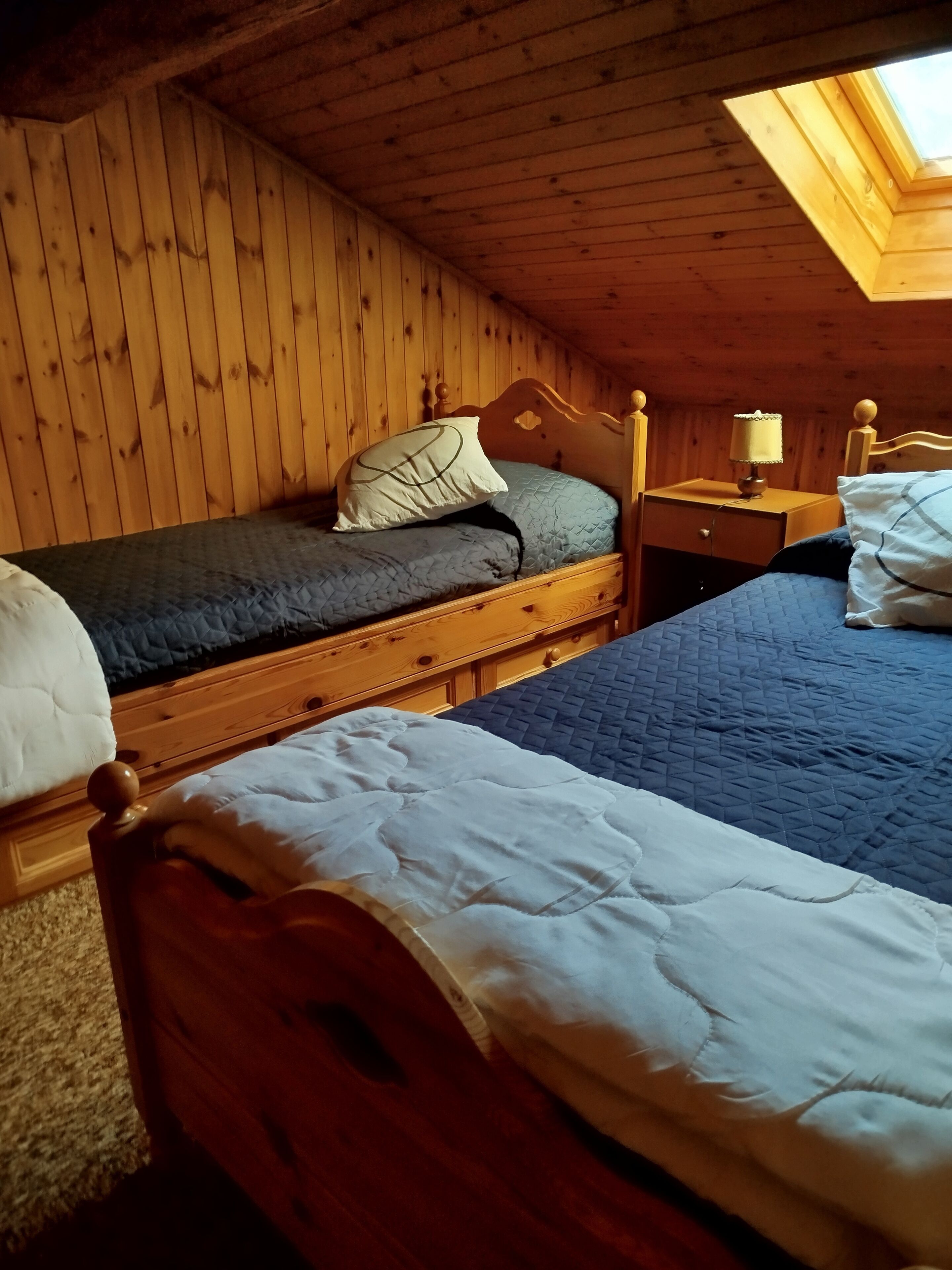 Une chambre mansardée chaleureuse, avec deux lits en bois, une lucarne et une petite table de chevet avec une lampe.