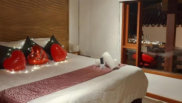 A hotel room with a large bed, a wooden headboard, a window with a view of the outside, and a bedside table with a remote control. - Vivelavilla suites VDL (Villa de Leyva)