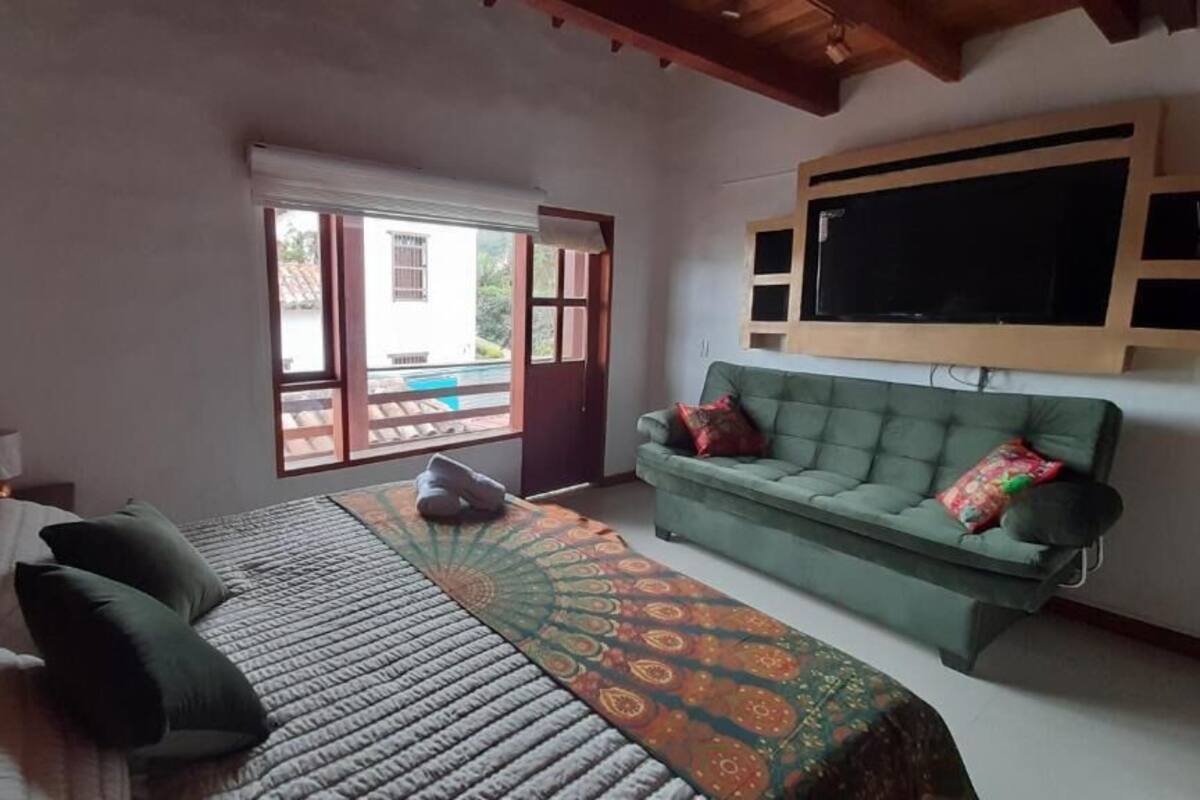 A bedroom with a bed, a green sofa, a TV mounted on the wall, and a window with a view of the outside.