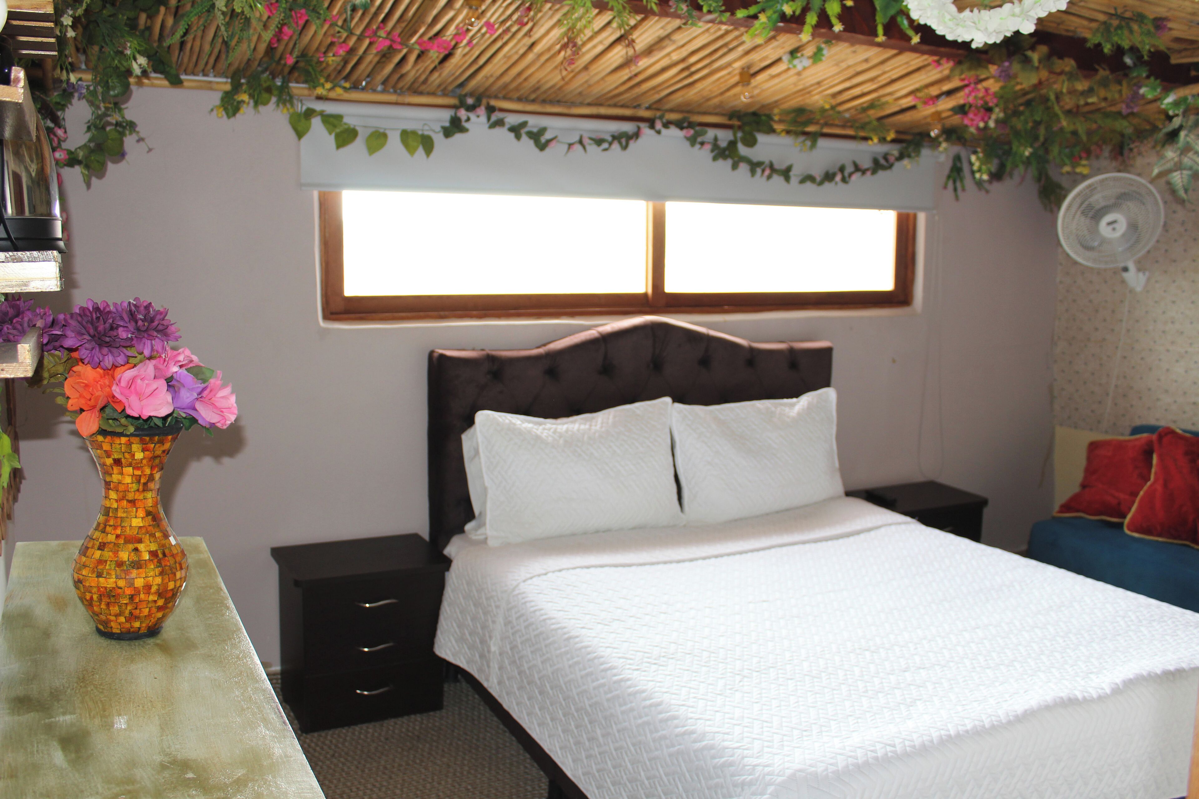 A bedroom with a bed, bedside tables, a nightstand, a vase with flowers, and a window.