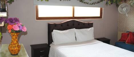 A bedroom with a bed, bedside tables, a nightstand, a vase with flowers, and a window.