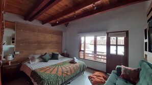 A bedroom with a wooden headboard, a bed with patterned bedding, a green sofa, and a window with a view of buildings. - Vivelavilla suites VDL (Villa de Leyva)