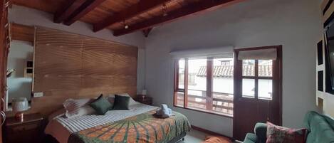 A bedroom with a wooden headboard, a bed with patterned bedding, a green sofa, and a window with a view of buildings.